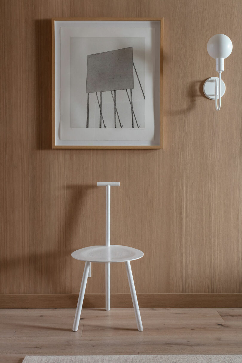 A minimalist wooden room has a framed abstract work of art, a modern white wall lights and a white chair with a unique backstream design.
