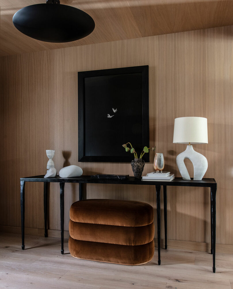 A modern console table with sculptural decor, a lamp and a vase sits against a wooden wall with a brown velvet east and a work of art framed with black.