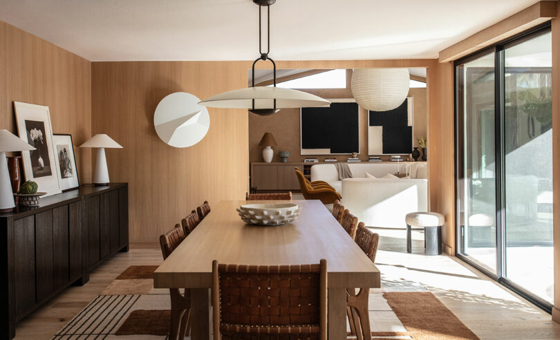 A modern dining room with wooden table, woven chairs, sideboards, decorative lamps and large sliding doors that get natural light.