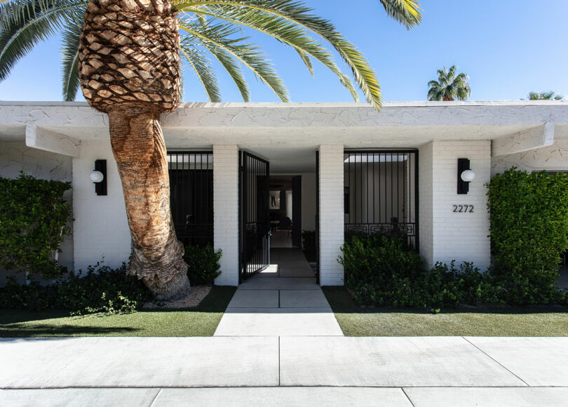 A modern, white brick building with black metal gates, a large palm and number 2272 at the entrance, surrounded by cut green.