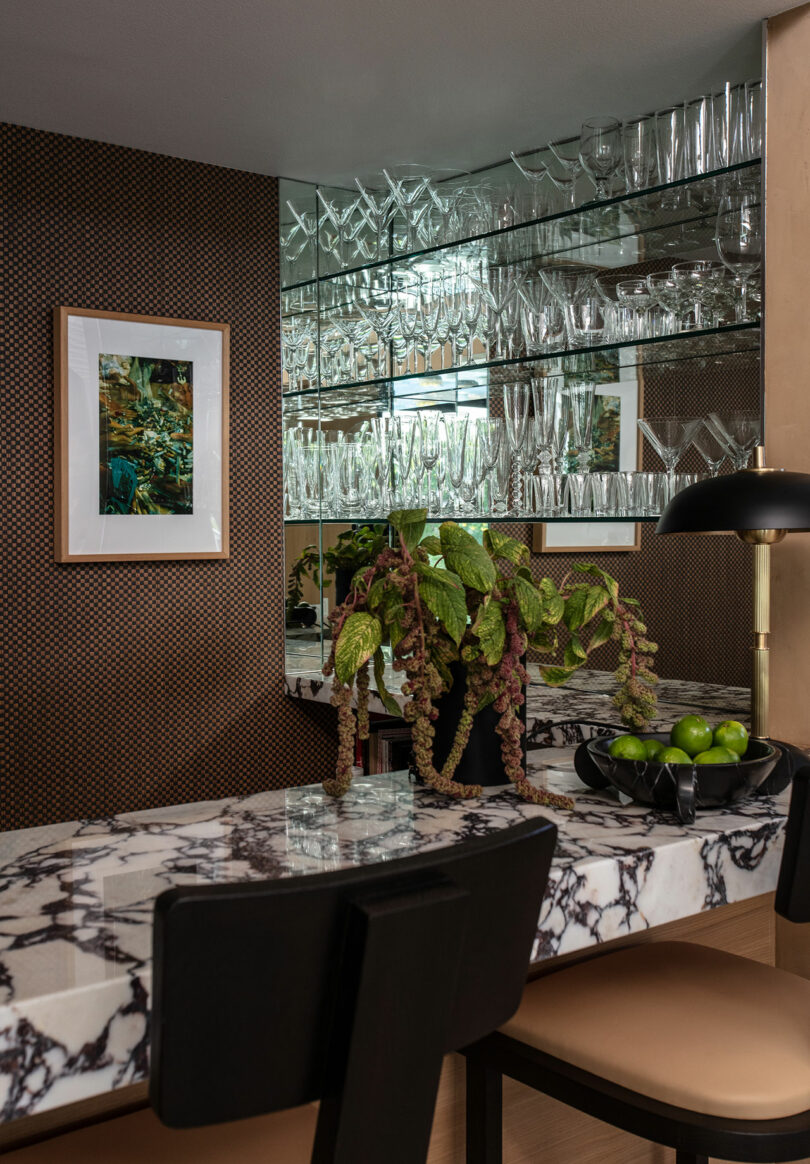 A marble dinner with black chairs stands in front of the mirror shelves filled with glassware, a leaf plant, a bowl with green apples and a black lamp. A framed picture hangs on the wall.