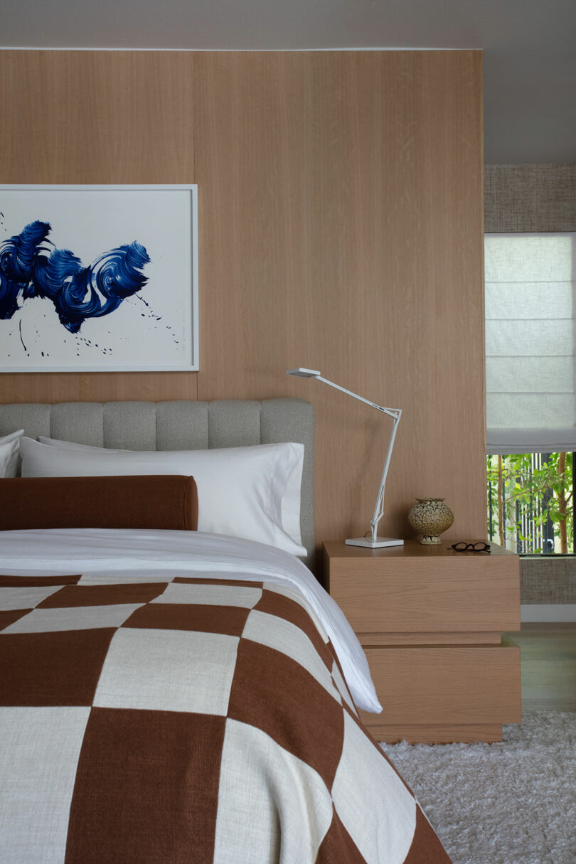 A modern bedroom with a light wooden accent wall, a gray, padded headboard, a bed with a brown and white checkered blanket and a bedside table with a lamp and decorative objects.