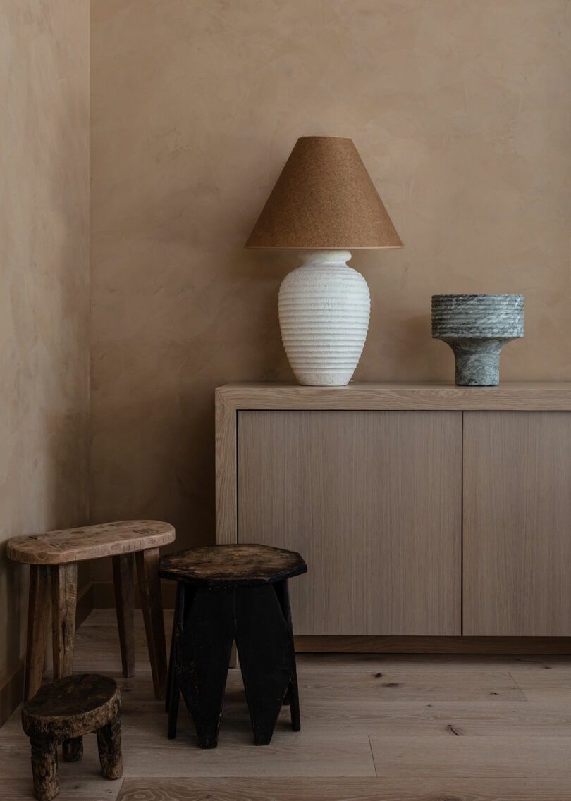 A minimalist room corner with a light wooden cabinet, a white lamp with a brown shadow, a green bowl and two rustic wooden hops on a wooden floor.