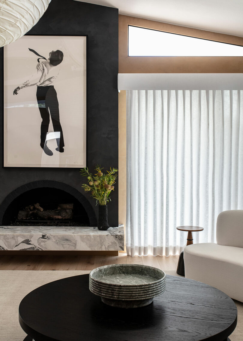 Modern living room with a black fireplace, abstract art over the fireplace, light curtains, a white armchair and a round dark coffee table with a ceramic bowl.