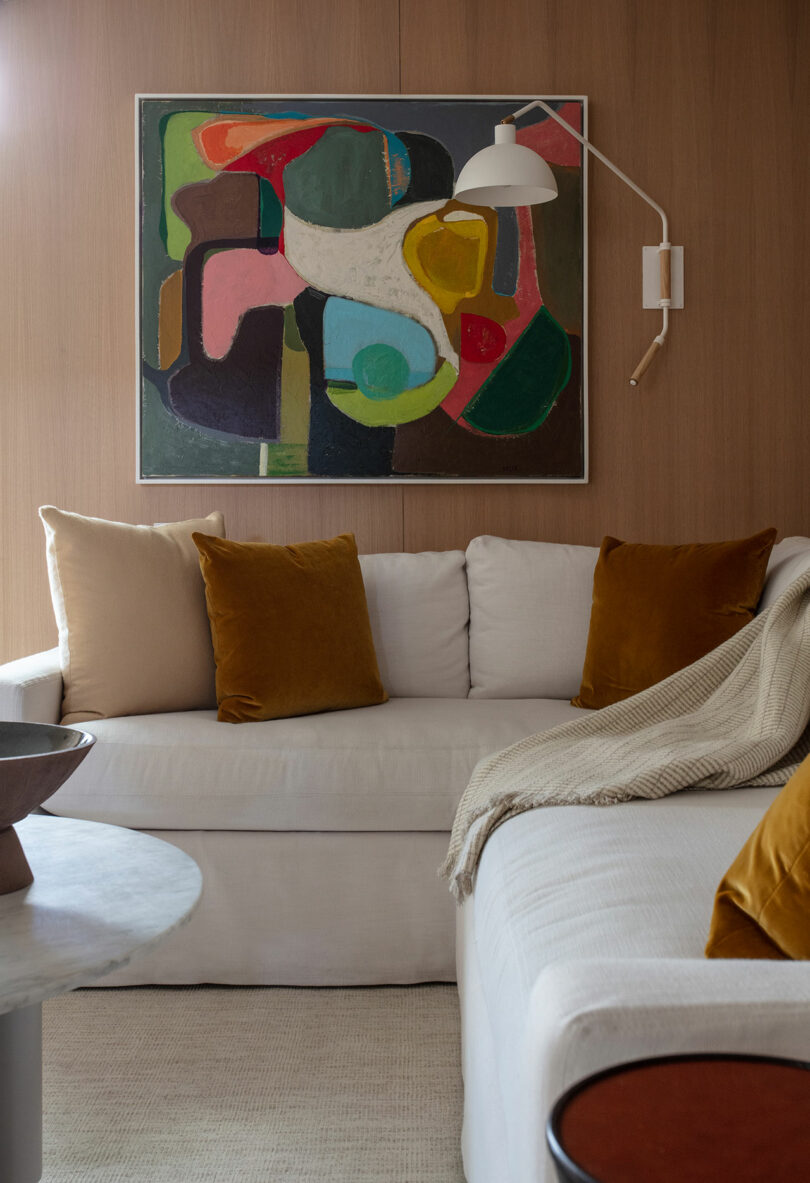 A modern living room with a white clippings, mustard yellow and beige pillows, a beige litter, an abstract wall art and a white wall lamp.