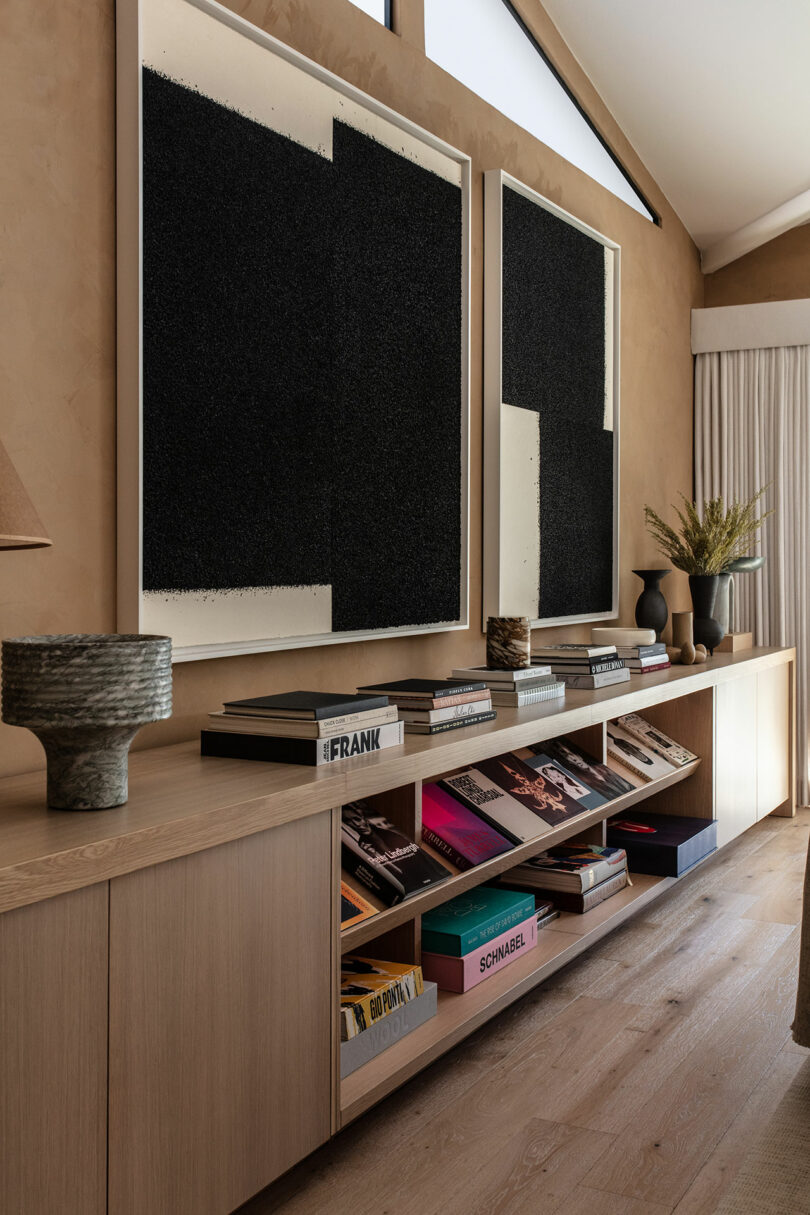 A modern living room with a long wooden sideboard, large abstract black works of art at the top, books and decorative objects that are arranged properly above and shelves below.