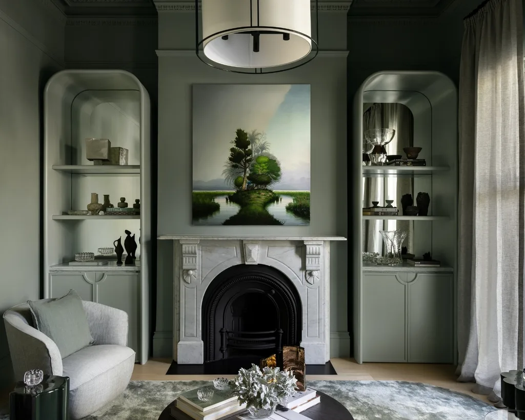 Elegant green living room with fireplace, modern art over it and shelves with decor. Cozy chair and gentle lighting beautify the room.