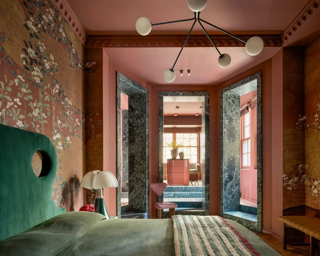 Cozy bedroom with flower wallpaper, green headboard, unique chandelier and view into a adjoining room with large windows. 