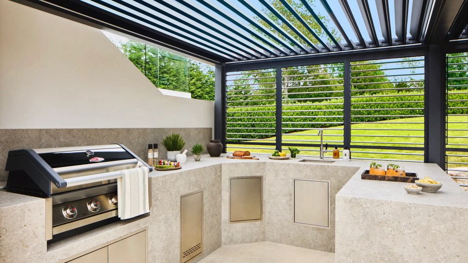 An outdoor kitchen with grill, sink and L-shaped counter under a louvered pergola overlooking a green lawn.