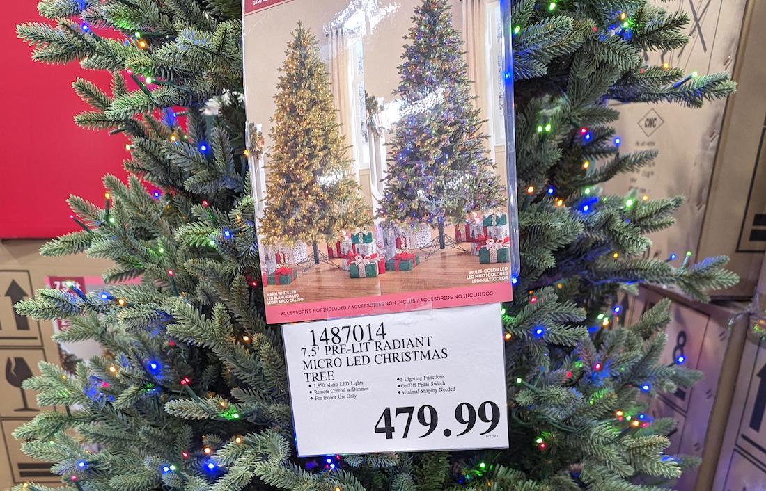 A 7.5-foot-tall pre-lit Christmas tree with multi-colored micro-LED lights is on display in one store. A sign below lists the price as $479.99, and a product photo shows decorated trees with gifts underneath.
