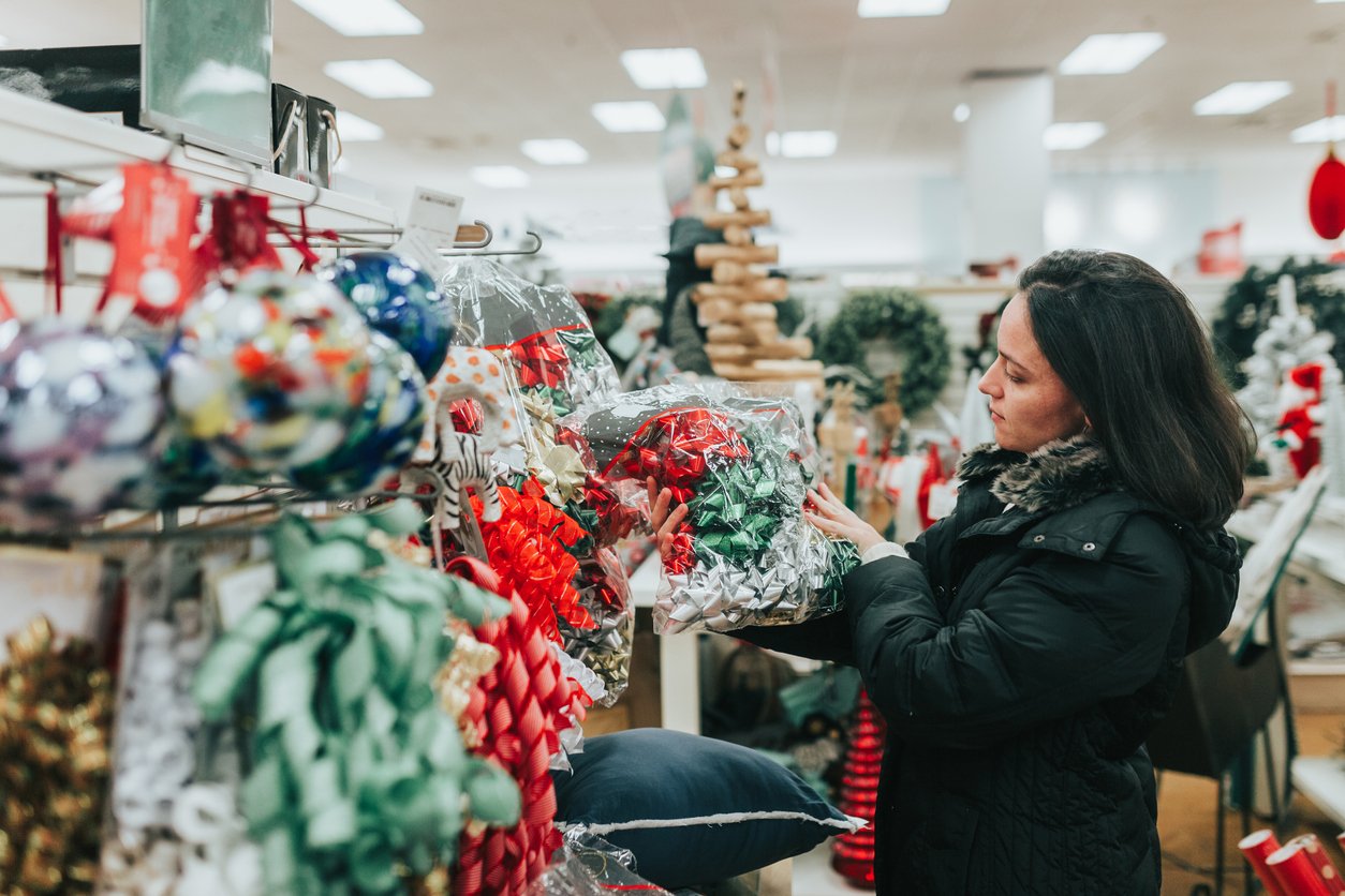 A woman in a black coat looks at a package of colorful gift bows in a store aisle decorated with Christmas items, including wreaths and a wooden Christmas tree.