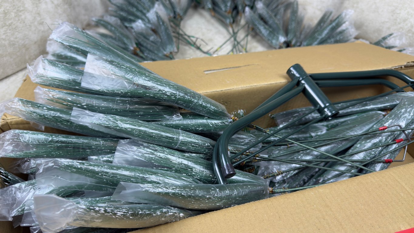 A cardboard box filled with artificial fir branches wrapped in plastic, next to a green metal wreath frame. More fir branches can be seen in the background.