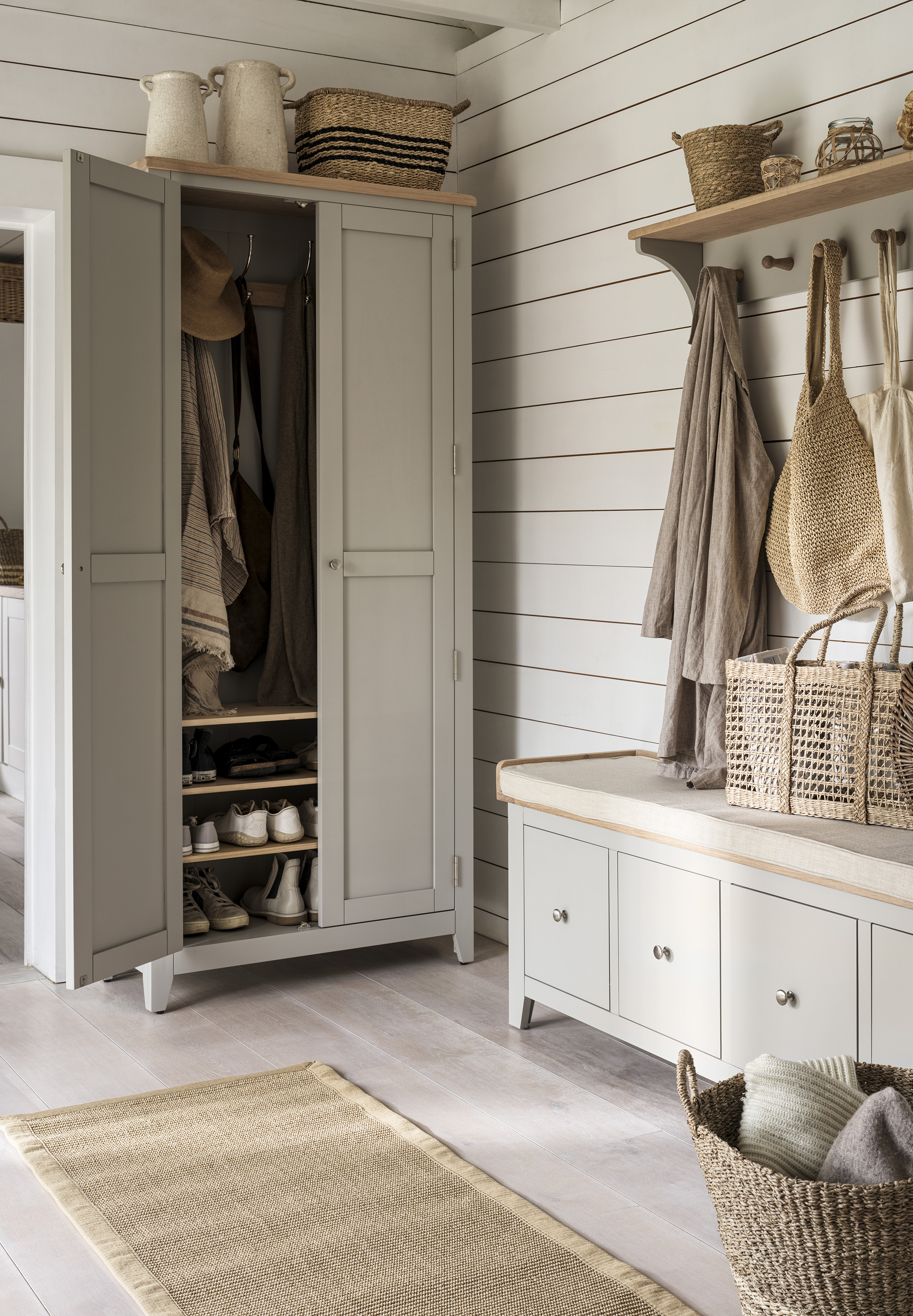 A gray-painted freestanding wardrobe in a white ship's entryway, alongside a matching shoe bench with drawer storage and a wall shelf with hanging coats
