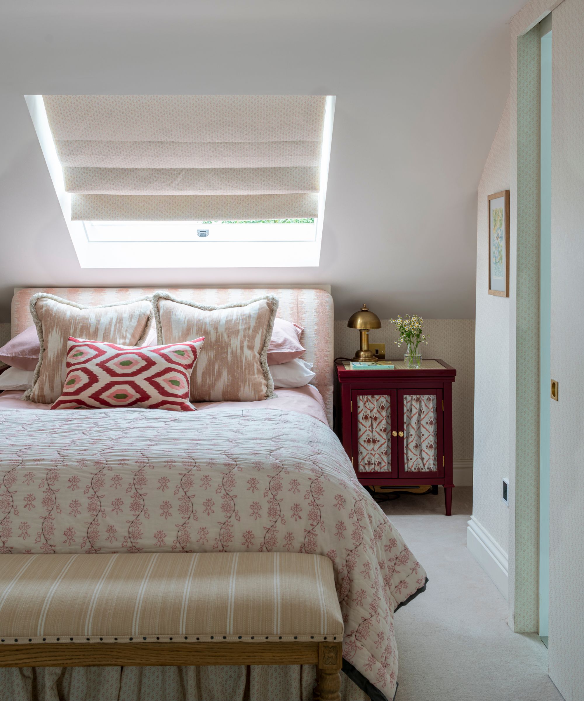 A white attic bedroom with a large double bed with pink and white patterned linens, pillows and headboard, a striped ottoman, a brown bedside table with a gold lamp and a white blind on a slanted window above the bed.