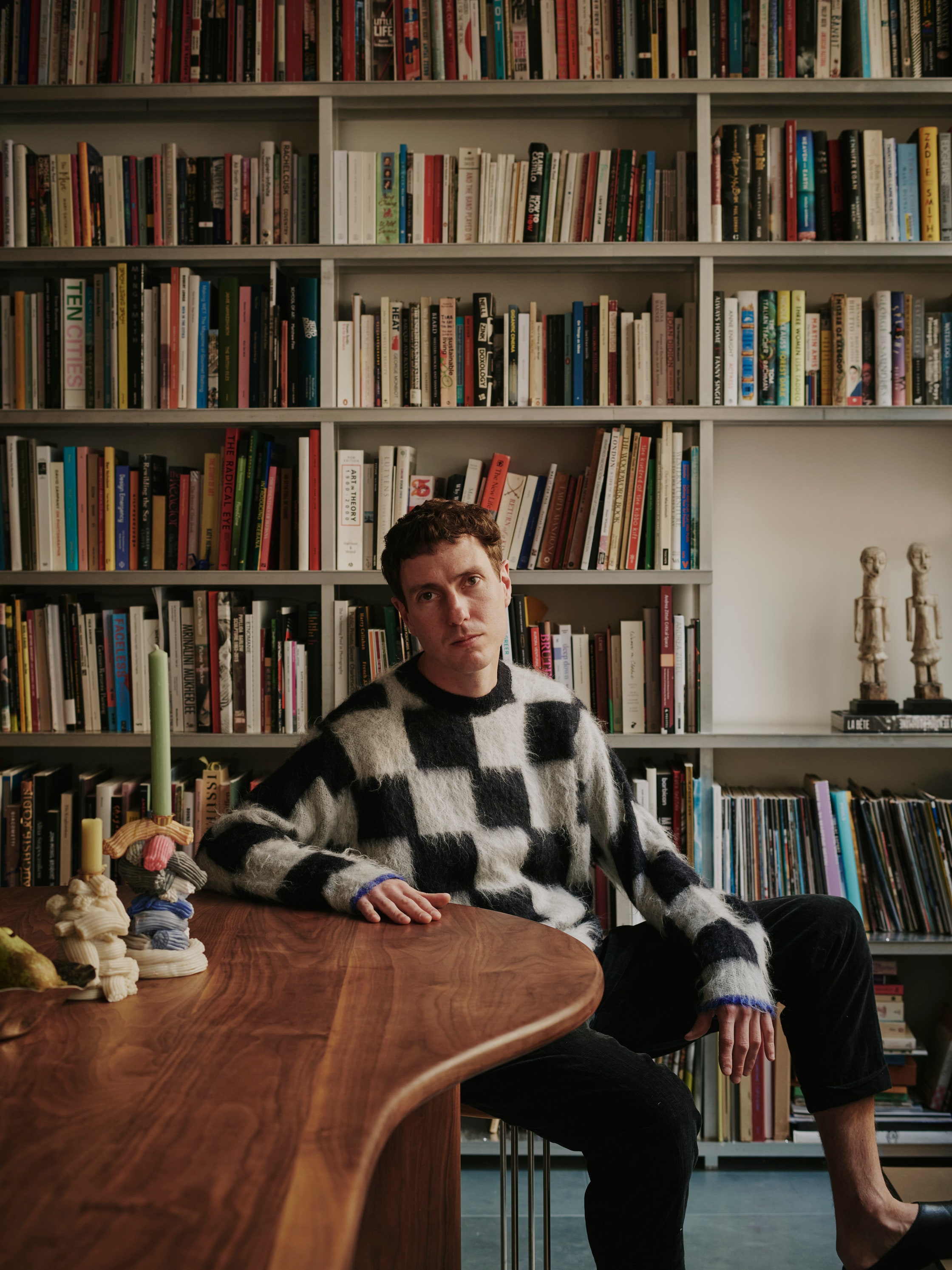 A young man wearing a checkered black and white sweater, black pants, and black leather pumps sits at a wavy wooden table next to soft candelabra in shades of blue, cream, pink, and yellow in front of a bookshelf full of tomes.