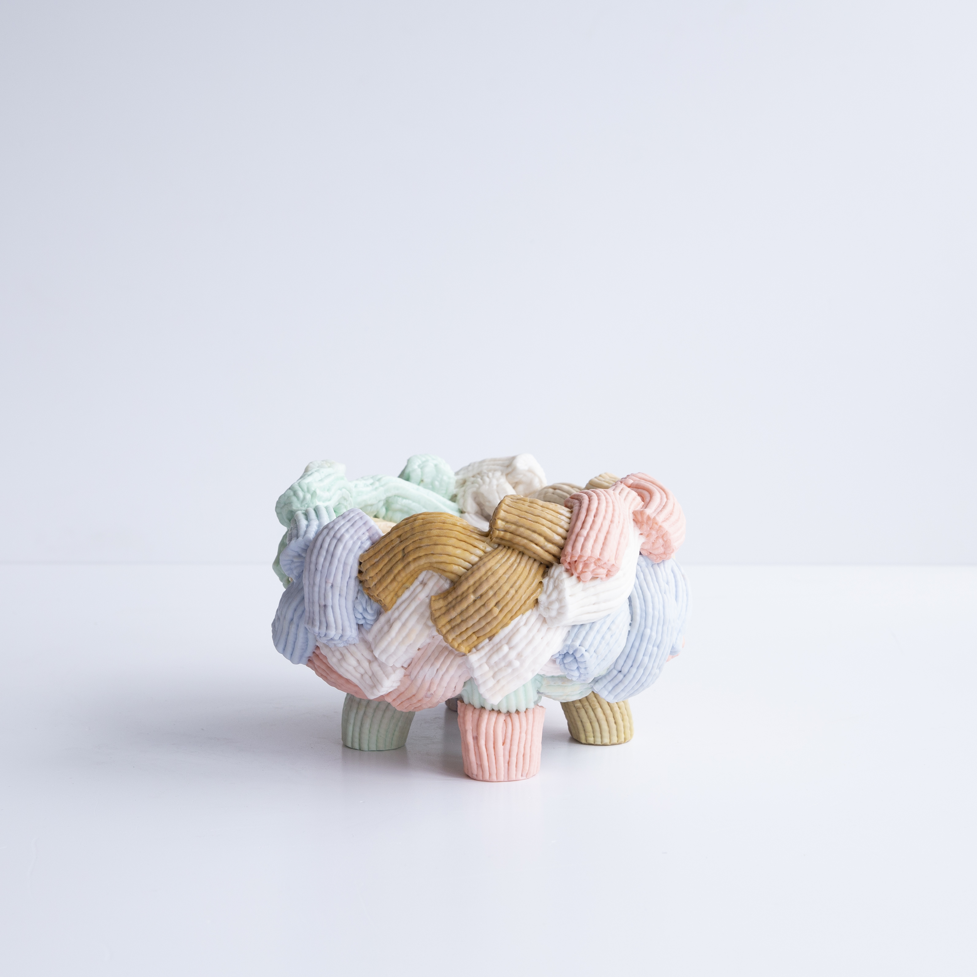 In a white room stands a pastel-colored vase with legs that consists of several surrounding plastic fragments that look like glaze.