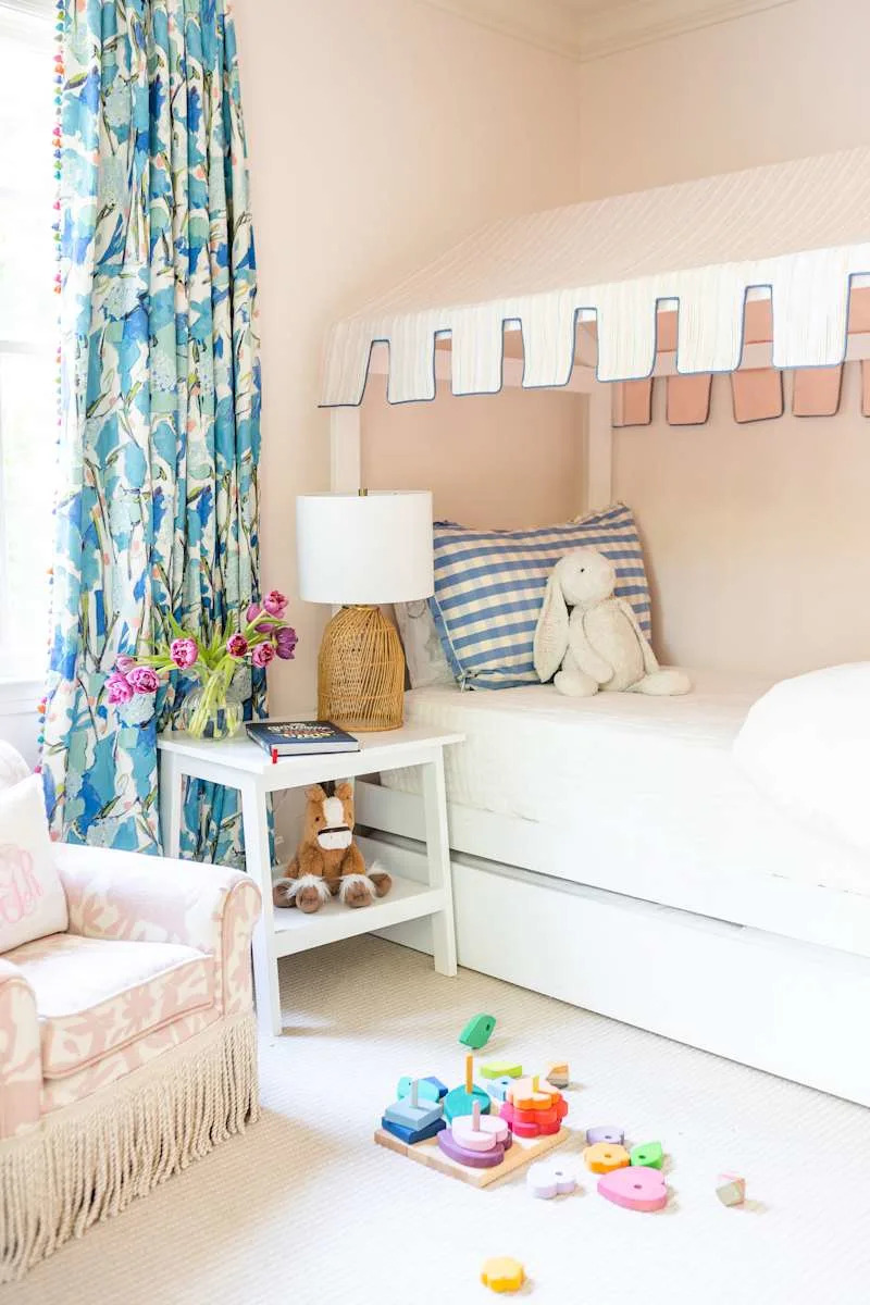 Brightly decorated children's room with a bed with a striped canopy, colorful curtains, a white side table with a lamp and toys on the floor.