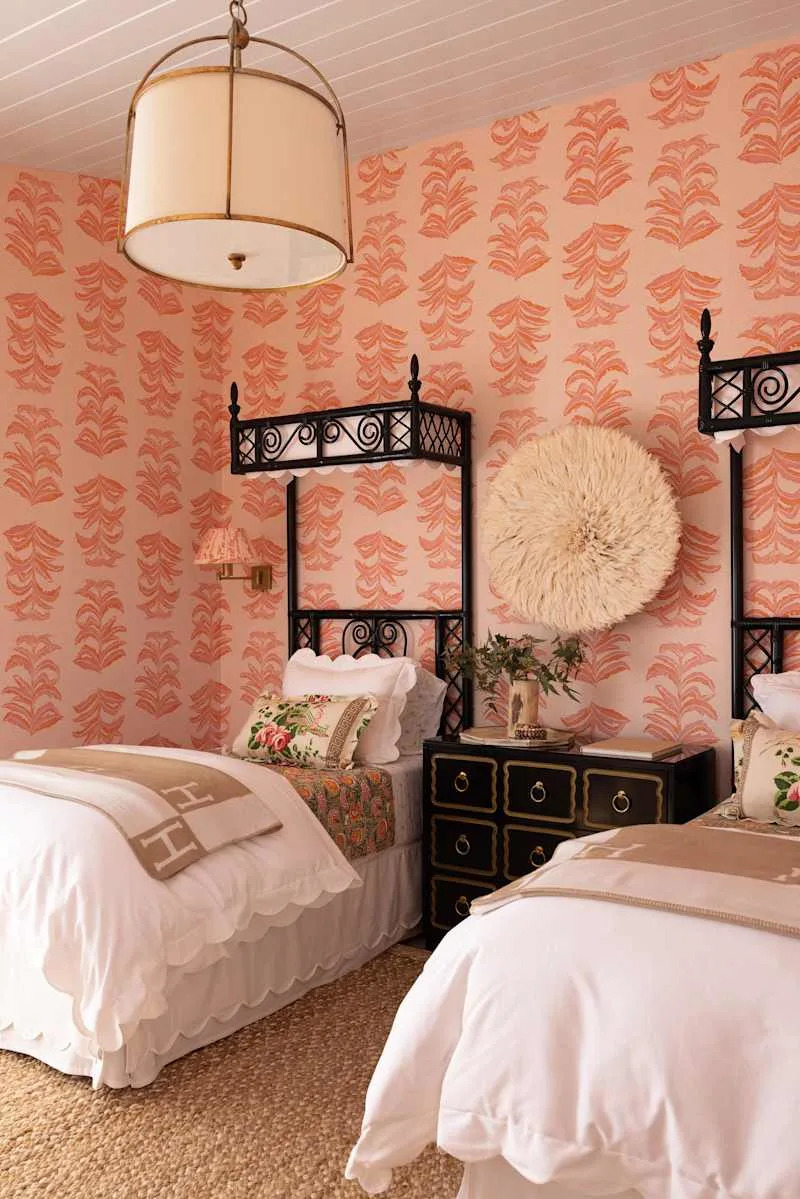 Two single beds with white linens and decorative pillows in a stylish bedroom with coral-patterned wallpaper, a black bedside table and a woven rug.