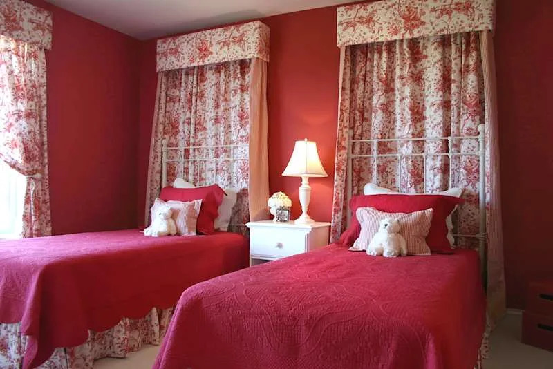 Two single beds with red quilts and decorative pillows, white bedside table with lamp and floral curtains.