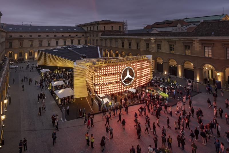 A large crowd enters an illuminated museum entrance with the Mercades logo in the center and yellow lights simulating the infamous car's grille.