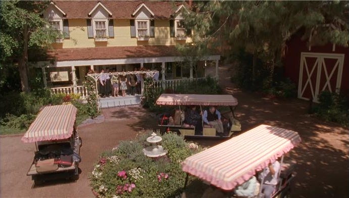 A scene depicting a group of people in front of a large yellow house with a porch surrounded by flowering gardens and golf carts
