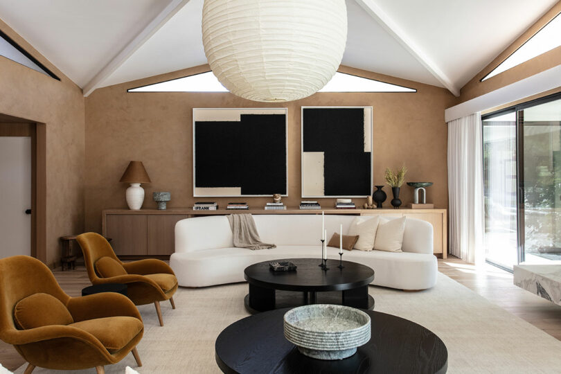 Modern living room with a white curved sofa, two mustard chairs, black round coffee tables, large paper lantern light, abstract wall art and natural light from sliding doors.