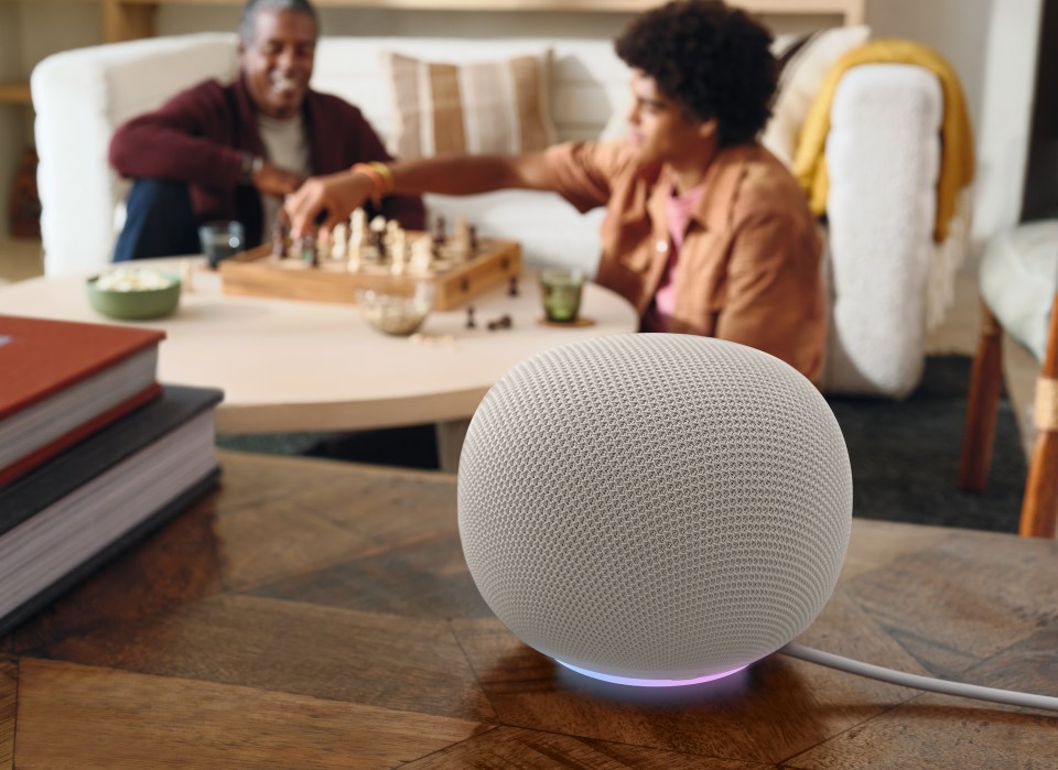 An intelligent loudspeaker with a shining base sits on a wooden table, while two people play chess in the background.