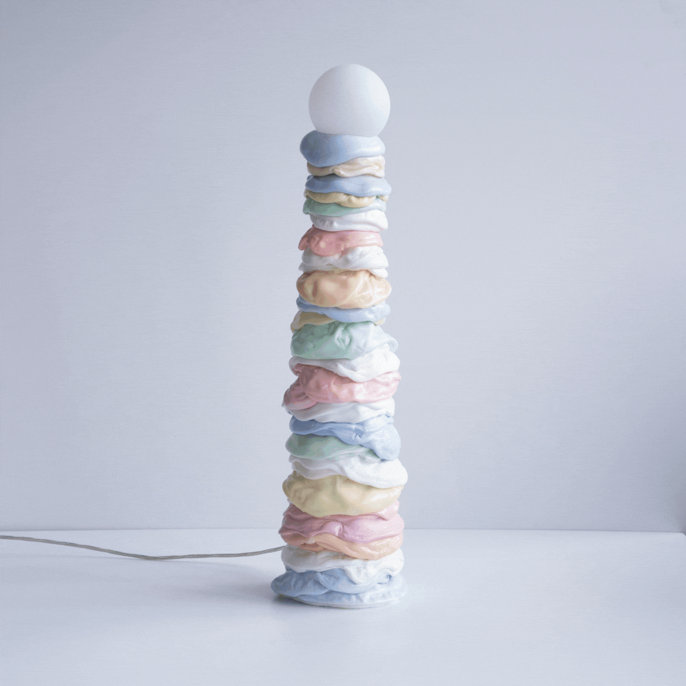 A squishy-looking lamp made of pastel-colored rings of melted plastic with a round light bulb on top stands in a white room as the light turns on and off intermittently.