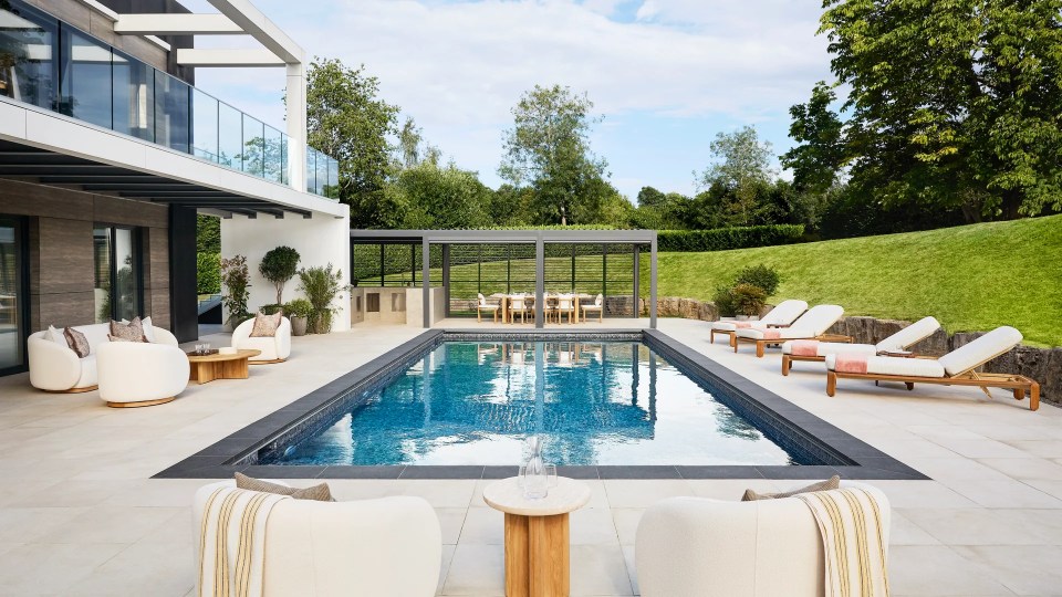 A modern house exterior with a swimming pool, outdoor lounge and dining area surrounded by a lush green hill.