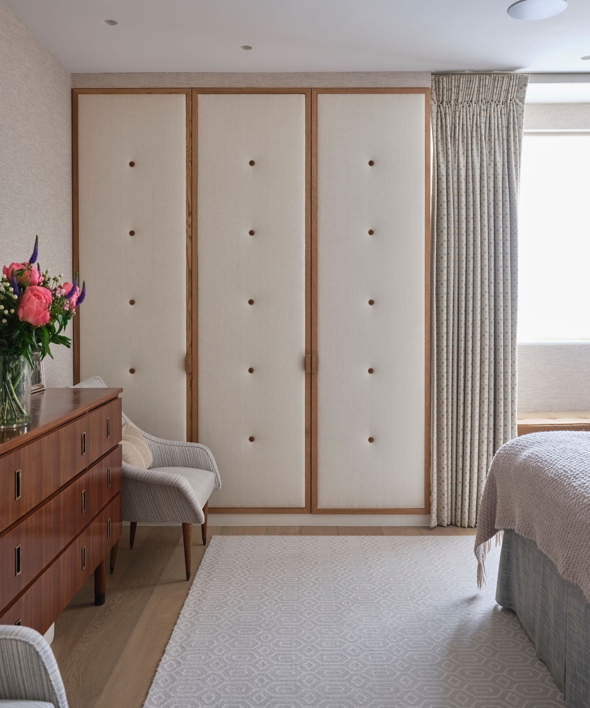 A cream and wood upholstered closet in a bedroom with a cream rug, a wooden sideboard with a vase of bright pink flowers and a gray chair.