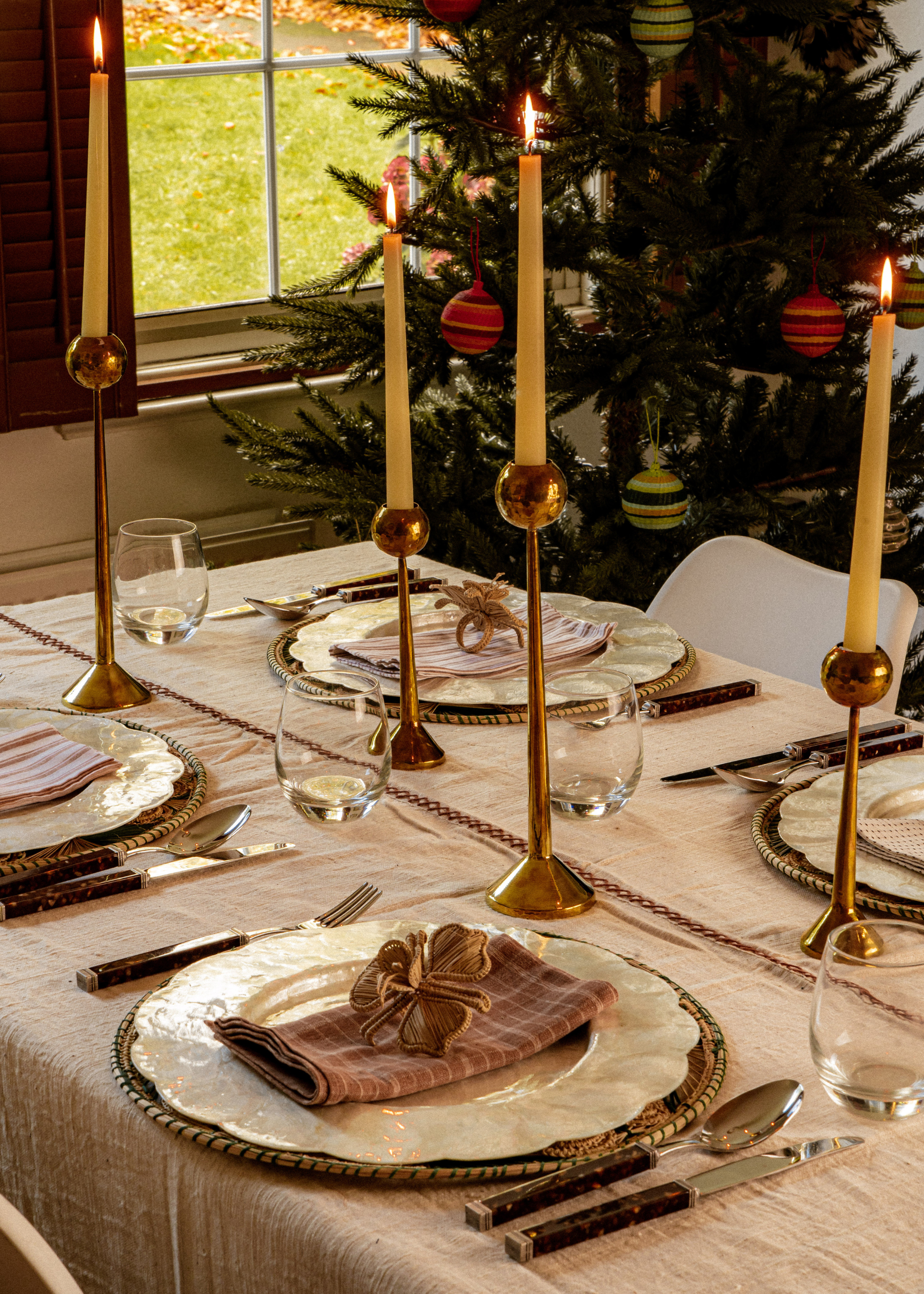 A dining table with plates, napkins, cutlery, glasses and candle holders next to a Christmas tree