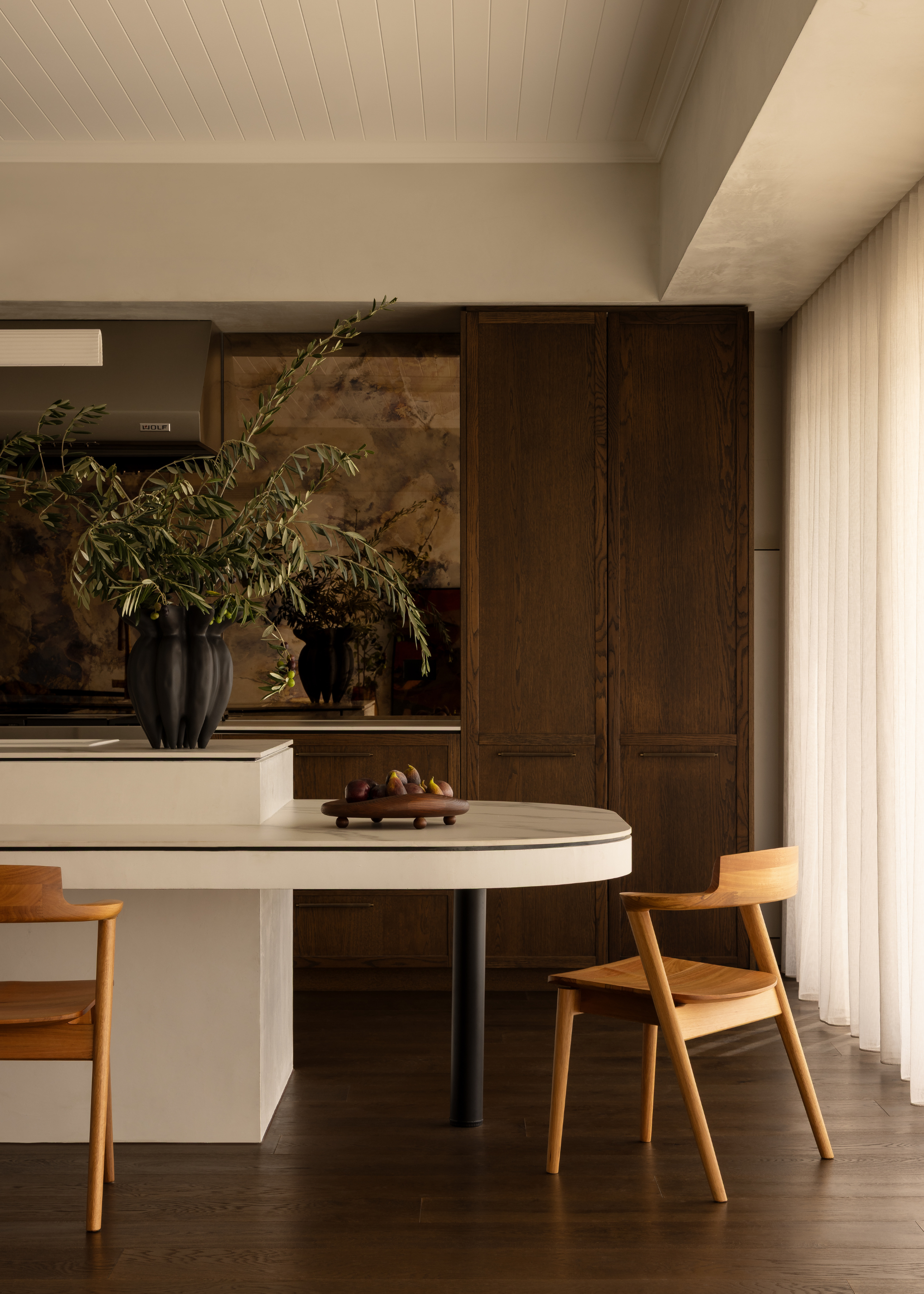 An open kitchen with a white island and annex next to wooden chairs
