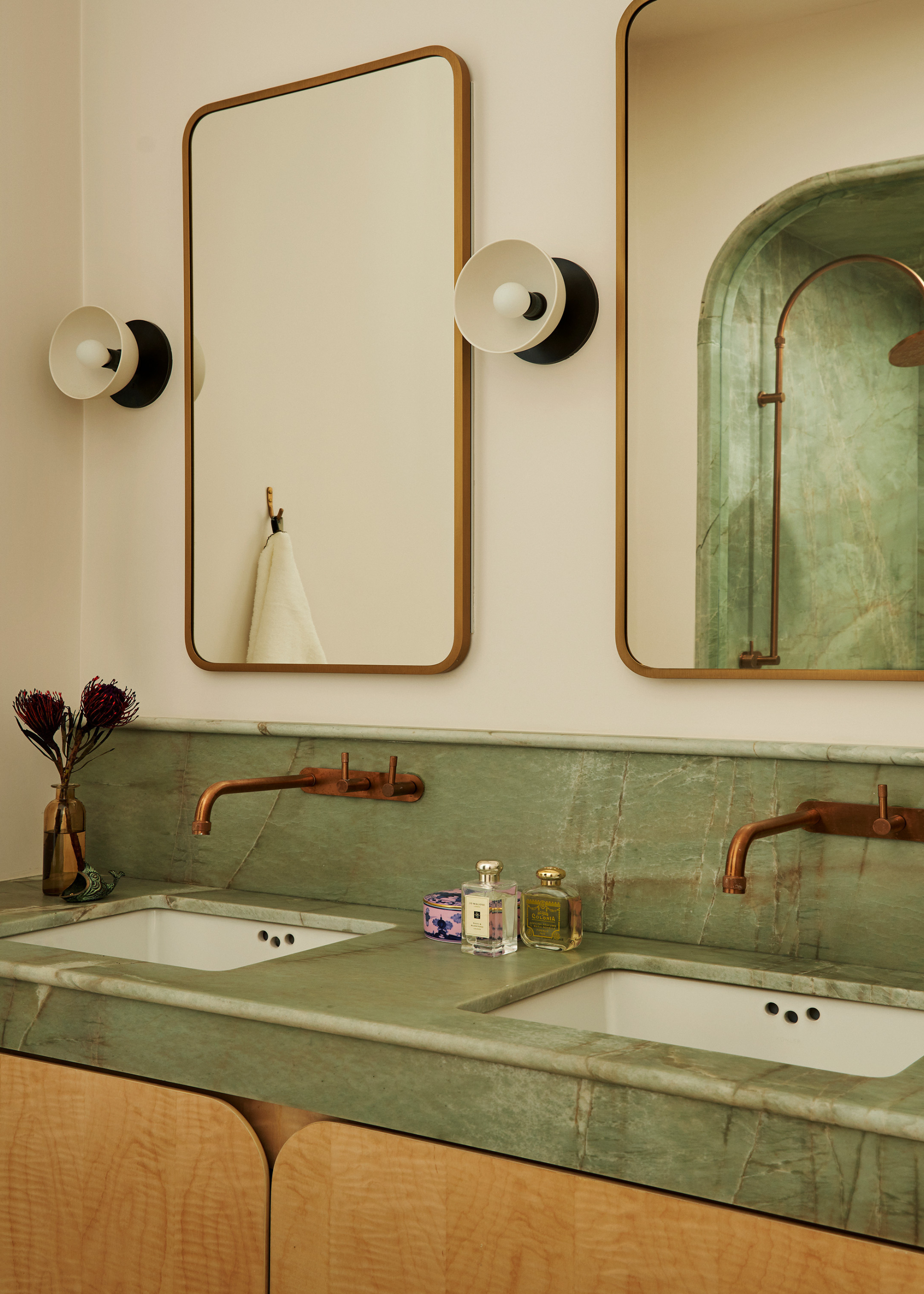 A bathroom with his-and-hers stations, a green marble countertop, gold-rimmed mirrors, black-and-white sconces, brass hardware, and wooden cabinets