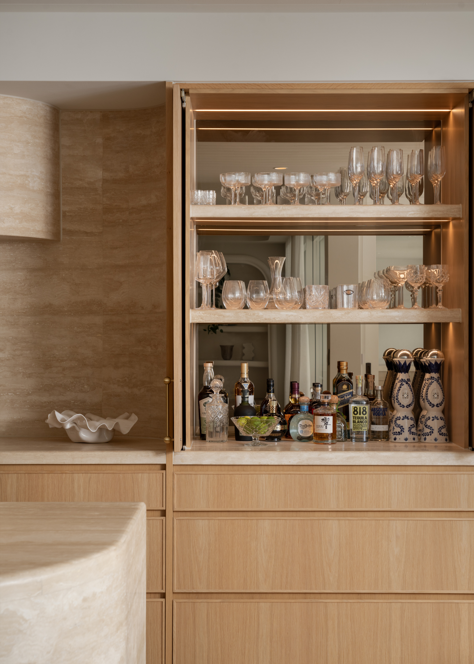 A wooden home bar with a mirror next to a kitchen with a minimalist marble backsplash