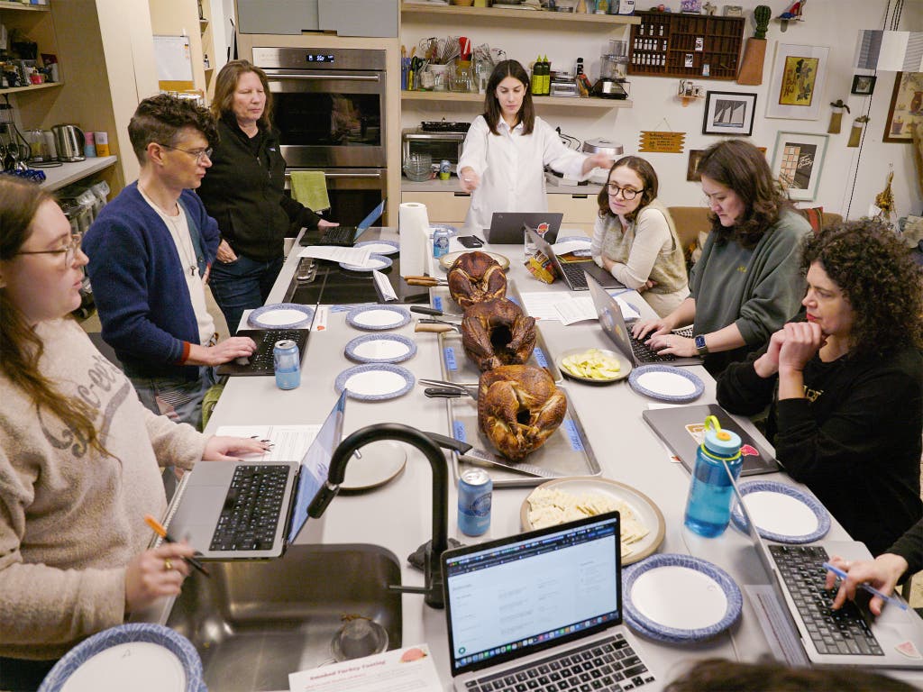 A group of people gather around a long table in a test kitchen to evaluate three whole smoked turkeys. There are laptops, paper plates and drinks available for tasting around the turkeys.