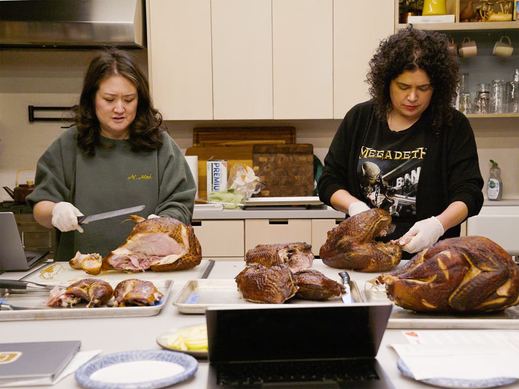 Two people wearing gloves stand side by side at a counter, carving several smoked turkeys onto sheet pans.