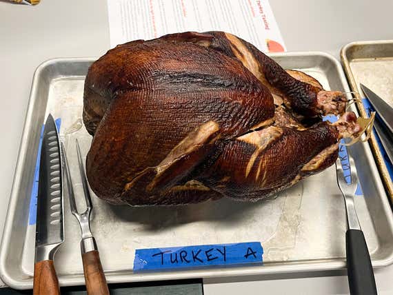 A whole smoked turkey lies on a sheet pan next to a carving knife and fork with blue tape written on it "Türkiye A" in the foreground.