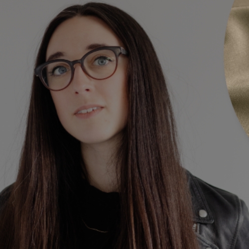 Image of a woman with brown hair and glasses in a black leather jacket standing against a gray background.