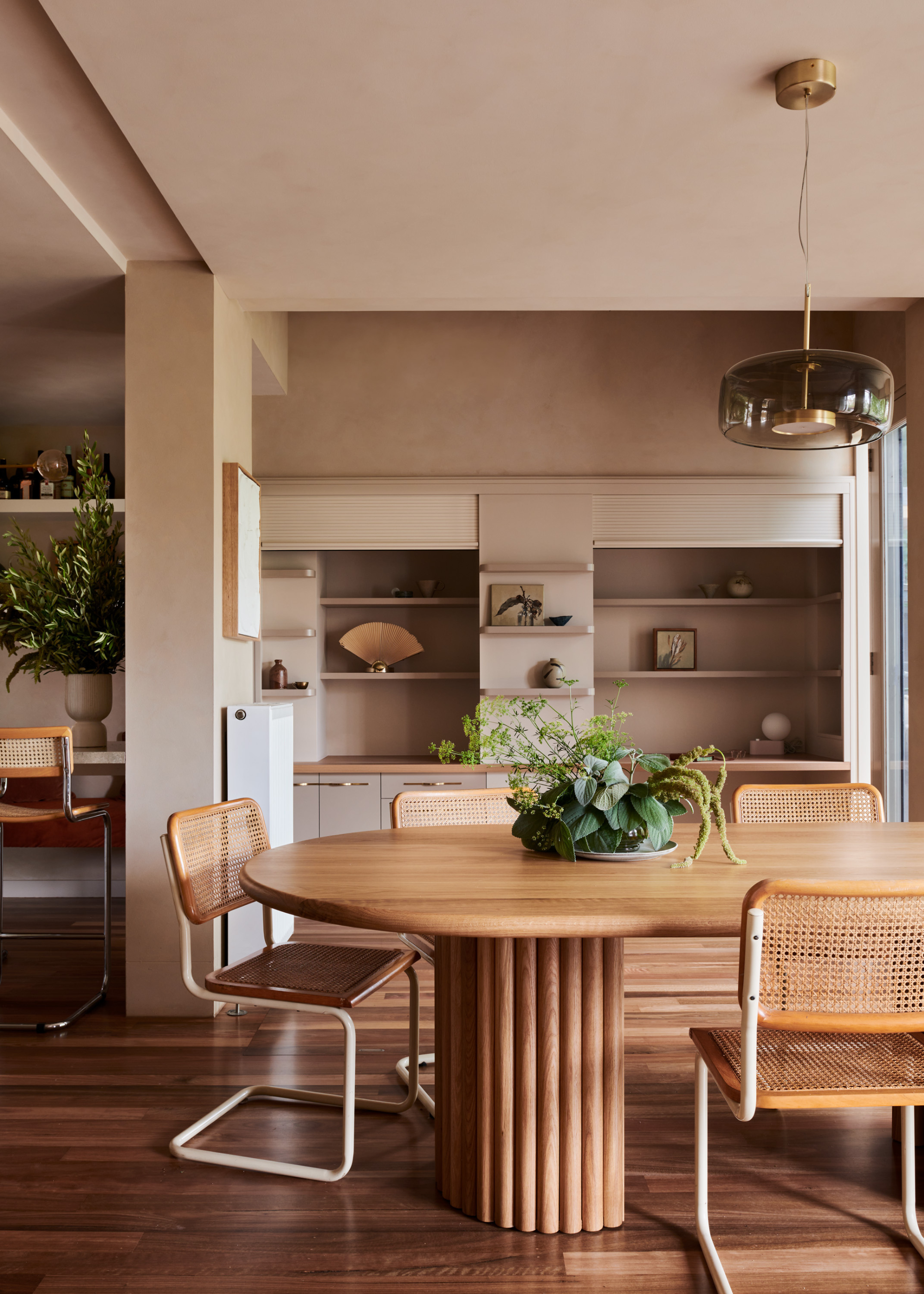 A dining room with beige walls, a glass pendant lamp, a wooden dining table with rattan-style dining chairs and a green centerpiece