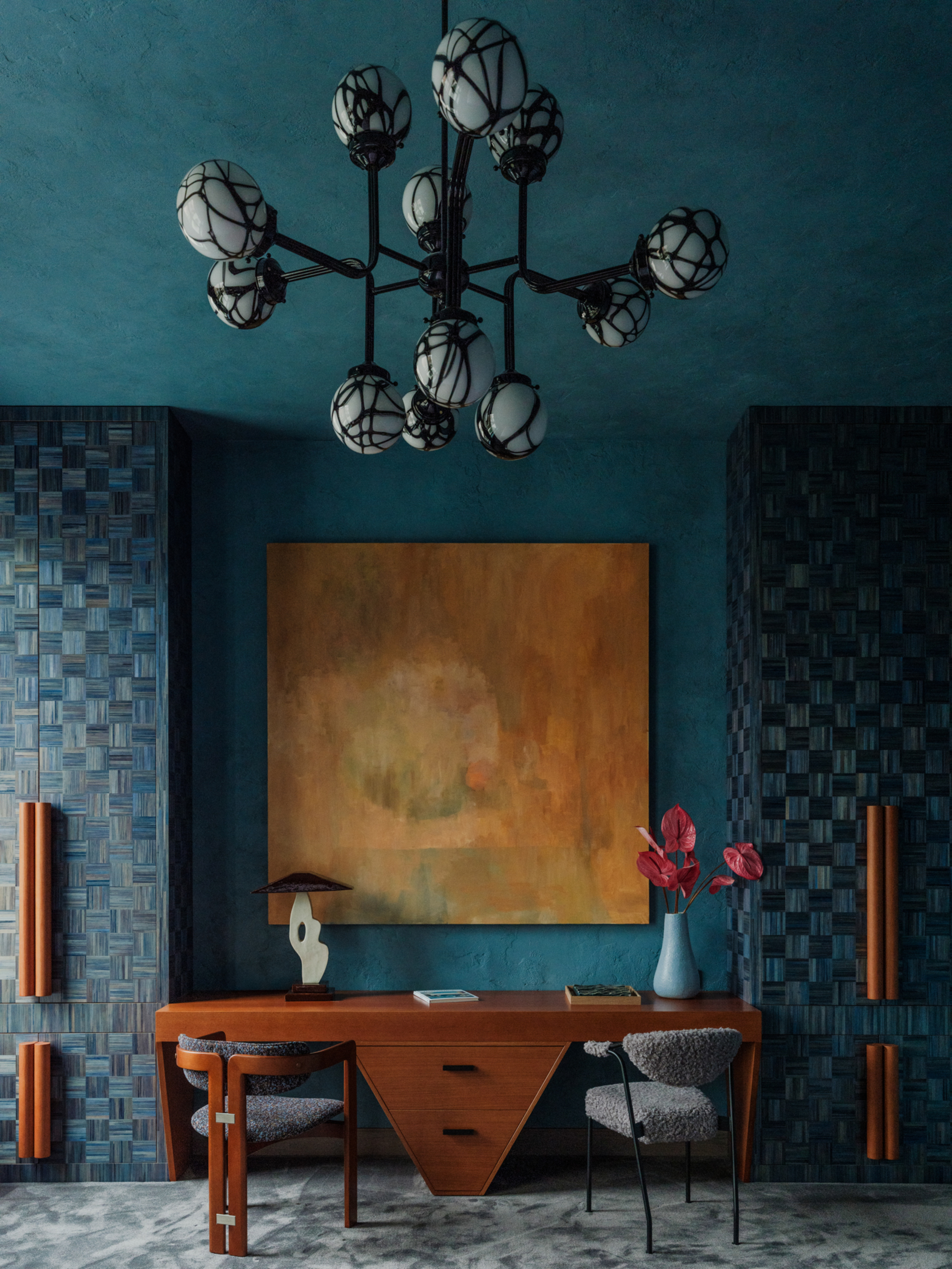 Image of a room with dark teal walls and ceiling and floor-to-ceiling cabinets made of blue-stained wood with a wicker pattern. There are gray carpeting, large orange handles on the cabinets, a large orange painting on the wall, and a geometric wooden desk.